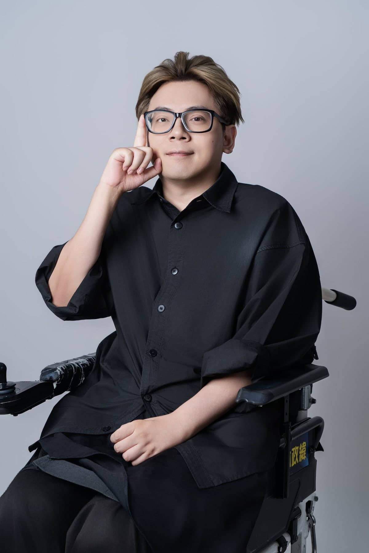 光合創學主理人光光（林政緯）的形象照：戴黑框眼鏡、穿黑色襯衫，坐在電動輪椅上，右手食指輕靠臉頰看著鏡頭，背景是淺灰色棚拍背景